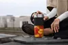 A person is holding a portable blender filled with fruit, sitting outdoors on a concrete structure. Voir les images du produit