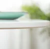 A close-up shot of the edge of a white round table with a teal object in the background, and blurred green foliage. Náhľad obrázku produktu