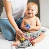 A baby is sitting on a white towel, holding a toy ball, with a person's hands nearby. Produktbild-Vorschau