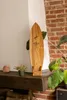 A wooden surfboard shaped product is displayed on a shelf in front of a brick wall, with plants and decorative rocks. Produktbild-Vorschau