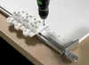 A drill is being used with a jig to drill holes in a white board, which is mounted on a metal rail. Produktbild-Vorschau