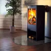 A modern black fireplace with a glass front sits on a glass hearth plate, next to a potted plant, in a room with white wooden walls. Produktbild-Vorschau