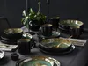 A table setting with a dinnerware set, including plates, bowls, and mugs, arranged on a gray tablecloth. A plant in a black vase and two black candle holders are also visible. Náhľad obrázku produktu
