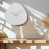 A white, organically shaped wall clock with wooden hands is mounted on a light-colored wall. Sunlight casts shadows across the wall and the clock. A wooden shelf is in the foreground with a decorative dried plant and a woven vase. Voir les images du produit