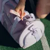 A hand is shown holding a pink strap with the word "Pusheen" on it, attached to a purple duffel bag. The bag is on a green surface with white lines, resembling a sports field. Produktbild-Vorschau