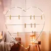 A heart-shaped wire frame decorated with fairy lights and small wooden clothespins, hanging in a softly lit room. Produktbild-Vorschau