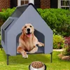 A golden retriever dog is lying in a gray dog house with a bowl of dog food in front of it, set in a garden with a brick house in the background. Produktbild-Vorschau