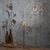 Two starburst-shaped LED lights are displayed against a textured gray wall. One light is on a stand in the foreground, and the other is hanging in the background. Dried reeds in brown glass bottles are to the left. Produktbild-Vorschau