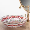 A decorative ceramic sink with a colorful floral pattern is shown on a wooden countertop, with a brass faucet and a round mirror in the background. Náhled obrázku produktu