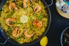 A close-up, top-down view of a paella dish in a pan, with shrimp, green beans, and lemon as garnishes. Other ingredients like garlic and mussels are visible in the background. Podgląd zdjęcia produktu