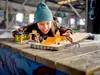 A young girl plays with a LEGO stunt park set, including a motorcycle and a spider-like vehicle, on a wooden surface with graffiti. Podgląd zdjęcia produktu