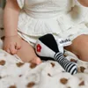 A baby's legs and hands are visible, holding a black and white rattle with a red flower design. The baby is wearing a white dress and is sitting on a patterned blanket. Produktbild-Vorschau