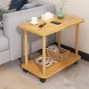 A small wooden side table with two shelves is placed next to a gray sofa in a living room setting. The table has items on it, including a remote control, a tissue box, a cup, a vase with a plant, and some magazines. Produktbild-Vorschau