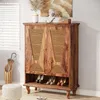 A wooden shoe cabinet with rattan doors, filled with shoes, in a room with blinds and a decorative stool. Anteprima dell’immagine prodotto