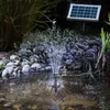 A solar-powered water fountain operates in a pond or garden water feature, with a solar panel visible in the background. Produktbild-Vorschau
