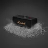 A black Marshall portable Bluetooth speaker sits on a pile of gray dust or powder, with some dust particles scattered around it. Produktbild-Vorschau