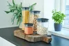 Four clear plastic food storage containers with black lids are arranged on a rustic wooden cutting board. The containers hold dry goods: spaghetti, beans, and lentils. A lush green plant is visible in the background, and a potted basil plant sits on a wind Náhľad obrázku produktu