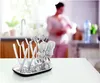 A baby bottle drying rack with bottles and nipples is in the foreground, with a blurred woman in the background in a kitchen setting. Produktbild-Vorschau