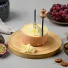 A cheese slicer is being used to cut a round of cheese on a wooden board. Other food items like grapes, passionfruit, and walnuts are visible in the background. Voir les images du produit