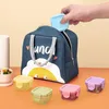 A hand is placing a small blue container into a blue lunch bag with a cartoon character design. Several other small colorful containers are arranged in front of the lunch bag. Produktbild-Vorschau