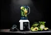 A blender filled with green vegetables sits on a wooden surface, with more vegetables arranged around it. Podgląd zdjęcia produktu
