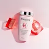 A bottle of Kérastase shampoo is displayed on a pink and white background, flanked by two red geometric decorative objects. Produktbild-Vorschau