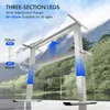 A white adjustable desk frame is shown in a natural outdoor setting with mountains and a lake in the background. The desk frame is shown in a partially extended state, with a blue arrow indicating upward movement. Produktbild-Vorschau