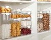A collection of clear plastic food storage containers filled with various dry goods, neatly stacked on white shelves. Produktbild-Vorschau