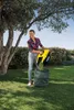 A woman is using a yellow and black garden tool to empty grass clippings into a green collection bag in a grassy yard. Produktbild-Vorschau