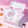 A white box filled with small white bags decorated with pink snowflakes and hearts, set against a pink, snowy background with snowflakes. Náhled obrázku produktu