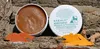 A tub of Keralit Undercover Pflegepaste is shown next to piles of orange and yellow powder, and some turmeric roots, all resting on a textured, bark-like surface. Produktbild-Vorschau