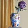 A decorative vase with an abstract pattern and a purple flower is placed on a wooden surface in front of a yellow curtain. Náhled obrázku produktu