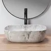A close-up shot of a marble sink with a black faucet, set against a light gray wall and a round mirror. Produktbild-Vorschau