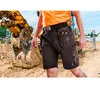 A construction worker wearing work shorts with tools in the pockets, standing in front of an excavator. Podgląd zdjęcia produktu