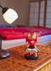 A Knuckles figurine wearing glasses and a smartwatch is placed on a woven surface next to a desk lamp, with a red bed in the background. Podgląd zdjęcia produktu