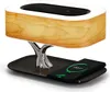 A modern bedside lamp with a wood-grain finish, a chrome tree-like stand, and a black base, with a smartphone charging on the base. Produktbild-Vorschau