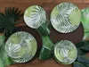 A set of plates and mugs with a tropical leaf pattern, arranged on a wooden surface with decorative leaves. Náhľad obrázku produktu