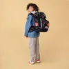 A young boy wearing a denim jacket and gray pants stands facing away from the camera, looking over his shoulder, with a large black backpack on. Anteprima dell’immagine prodotto