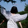 A person wearing angel wings and a halo stands outdoors, facing away from the camera, with trees and a blue sky in the background. Produktbild-Vorschau