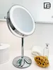 A chrome makeup mirror with LED lighting is displayed on a white surface, with a towel, a bottle of liquid, and a sponge in the background. A logo is visible in the upper right corner. Produktbild-Vorschau