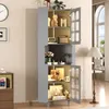 A tall, gray cabinet with glass doors stands in a room, filled with decorative items and illuminated by a floor lamp. Produktbild-Vorschau