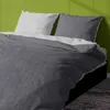 A close-up view of a bed with grey and light grey bedding, against a green wall. Produktbild-Vorschau