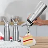 A hand is using a whipped cream dispenser to decorate a slice of cake, with two other dispensers and some berries on a table in the foreground. Produktbild-Vorschau