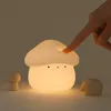 A hand is shown pressing the top of a glowing mushroom-shaped night light. A small wooden mushroom and a hedgehog figurine are also visible. Podgląd zdjęcia produktu