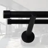 A close-up view of a black double curtain rod with a diagonal support bracket, set against a blurred background of a modern living room. Podgląd zdjęcia produktu