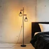 A floor lamp with two cage-style lampshades and wooden accents stands next to a bed in a bedroom with a concrete wall. Produktbild-Vorschau