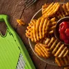 A close-up of waffle fries and a green mandoline slicer on a wooden table. Produktbild-Vorschau