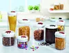 A collection of clear plastic food storage containers with white lids and red accents, filled with various dry goods like pasta, cereal, coffee beans, and candies, arranged on a white countertop in a kitchen setting. Produktbild-Vorschau