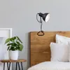 A black clip-on lamp is attached to the headboard of a wooden bed, next to a potted plant on a side table. Produktbild-Vorschau