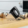 A black Bosch hand mixer is shown on a wooden countertop next to a metal mixing bowl and some bread. Produktbild-Vorschau